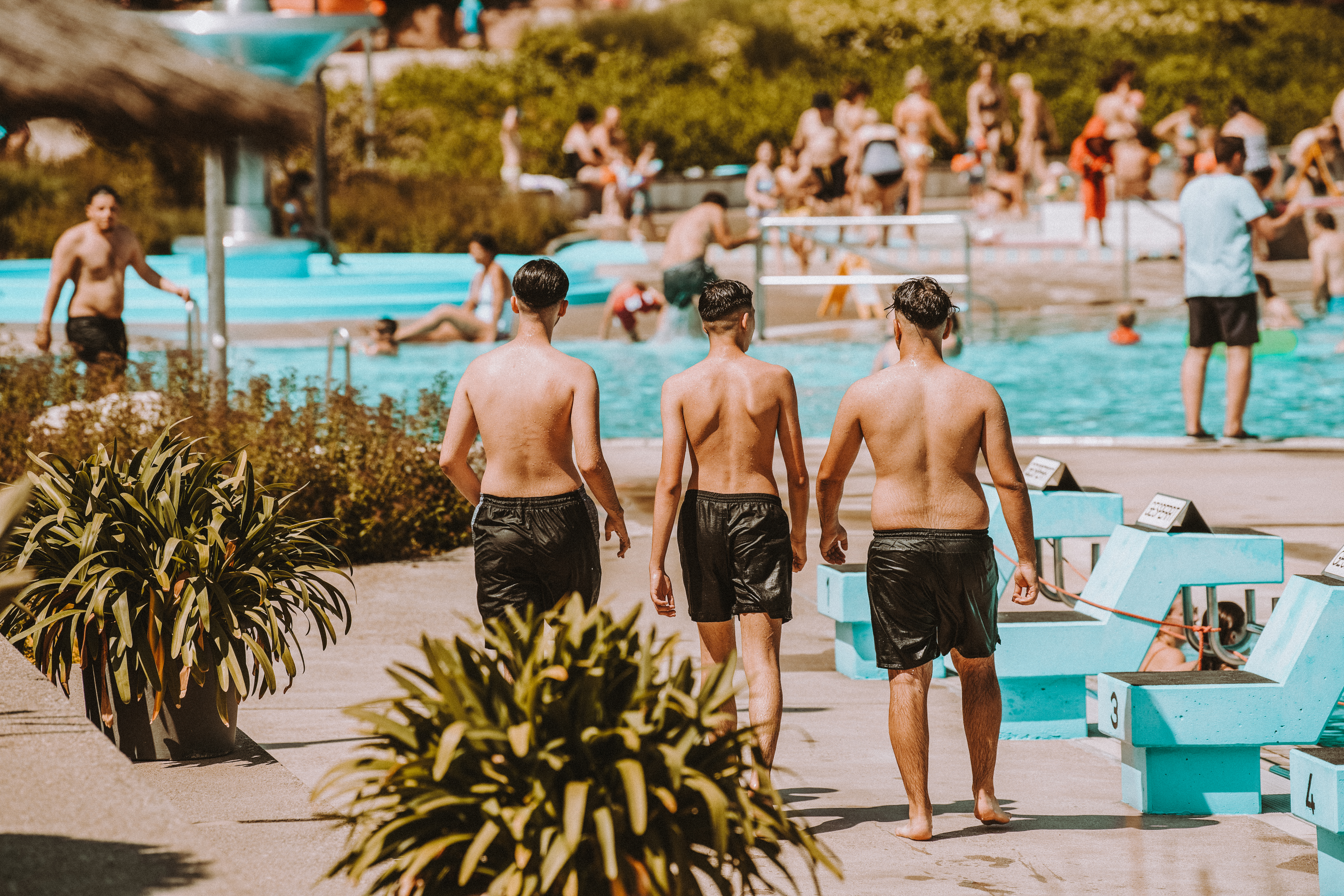  3 junge Männer gehen am Beckenrand entlang, im Hintergrund reger Badebetrieb 